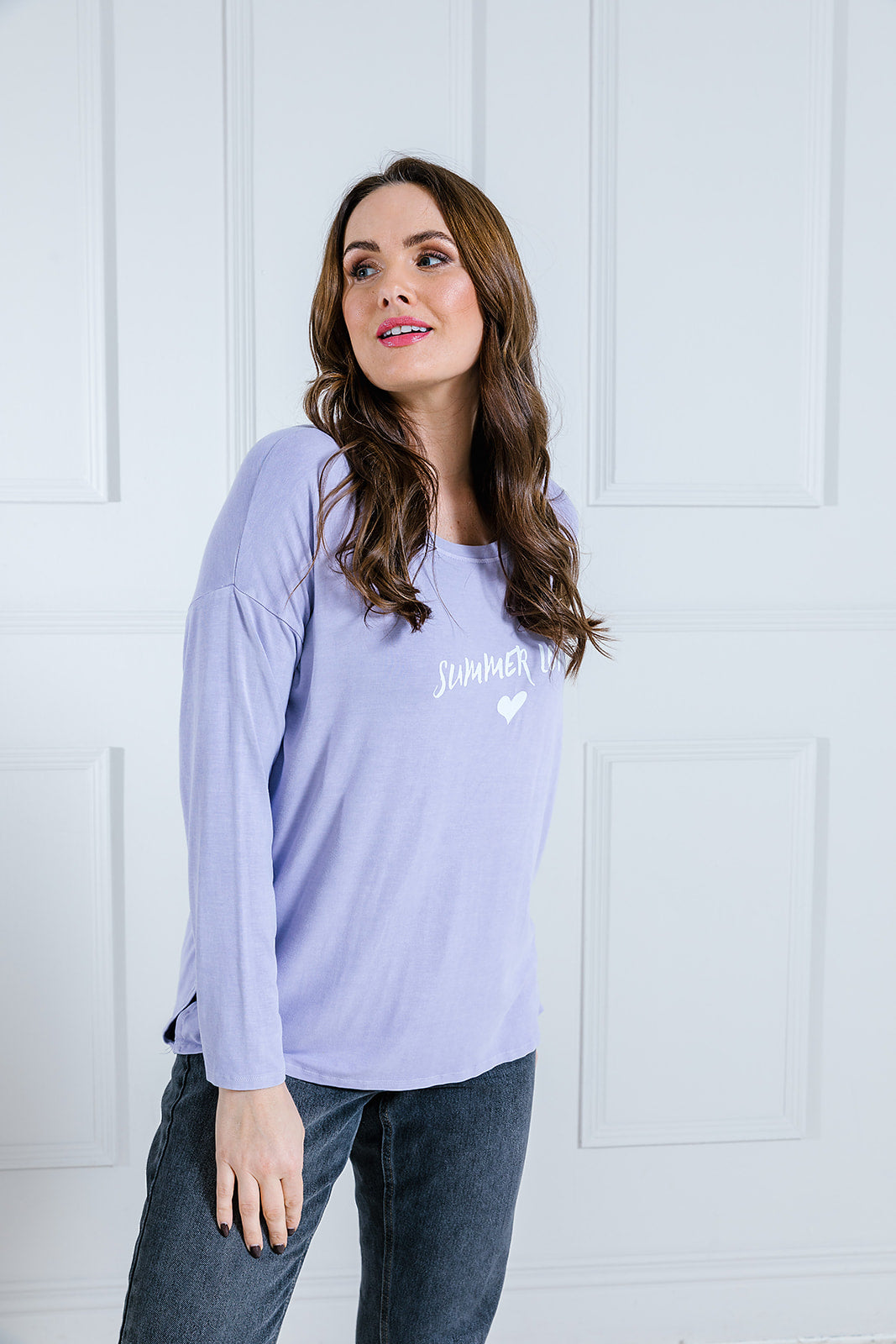 A woman wearing a long sleeved top with a scoop neck, printed with the phrase 'Summer Lovin'. The top is lilac in color and she is paired with dark pants.