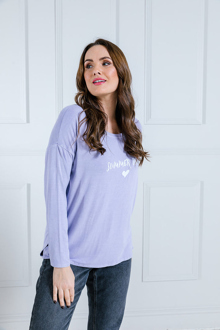A woman wearing a long sleeved top with a scoop neck, printed with the phrase 'Summer Lovin'. The top is lilac in color and she is paired with dark pants.